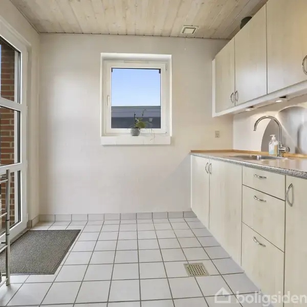 Køkkenområde med lyse skabe og skuffer langs en væg, vask og vandhane på bordpladen. Skohylde fyldt med sko ved dør, lyse fliser på gulvet, vindue med plante.