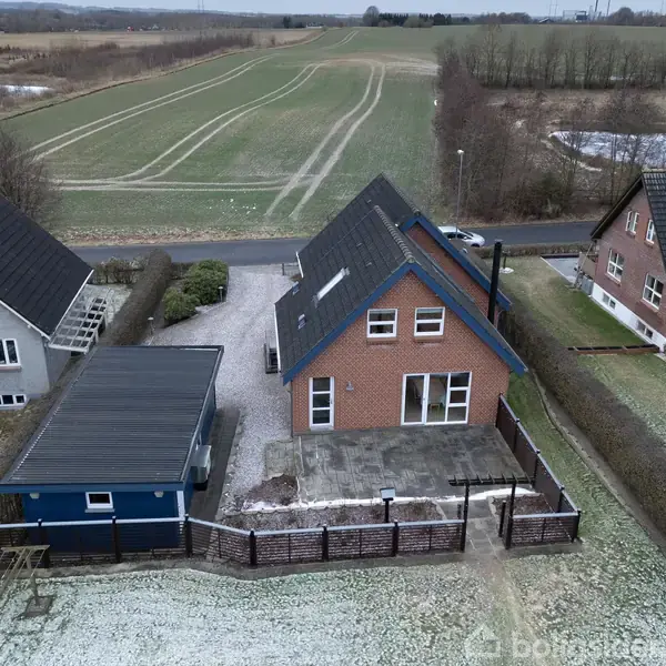 Et rødt hus med gråt tag står midt i en snedækket have. Bag huset ses en åben mark og en vej. Andre huse ligger tæt ved siden af.