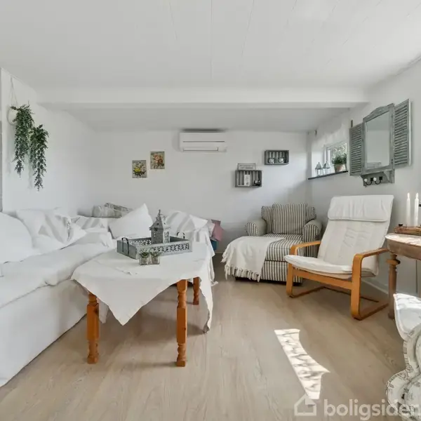 Stue med hvide sofaer og pyntepuder, står stille i et lyst rum med trægulv, hvor planter og dekorationer tilføjer hygge. Store vinduer lader naturligt lys ind.