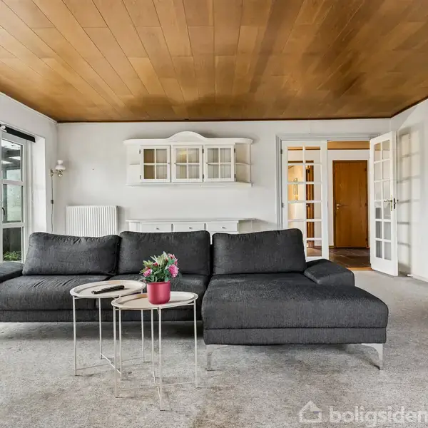 En grå sofa står i en stue med træloft, flisegulv og hvide vægge. Ved siden af sofaen står to små borde med en rød blomsterkrukke. I baggrunden ses glasdøre.