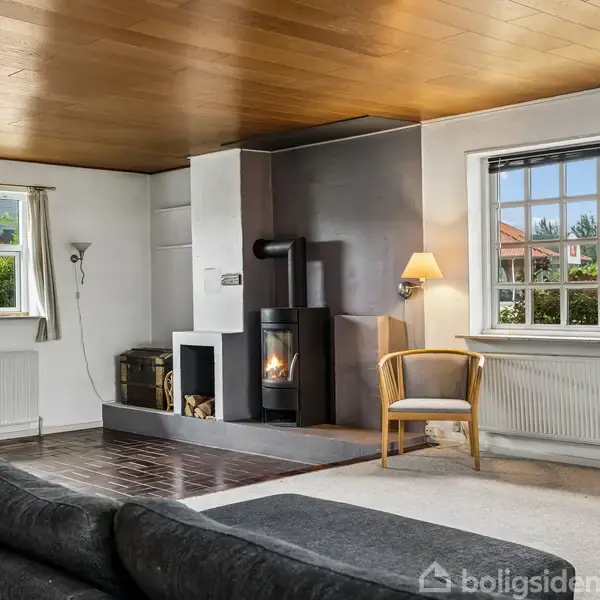 Sort sofa står i en stue ved siden af en brændeovn, der er i brug. Vinduet til højre viser en grøn have, og en stol står ved siden af brændeovnen.
