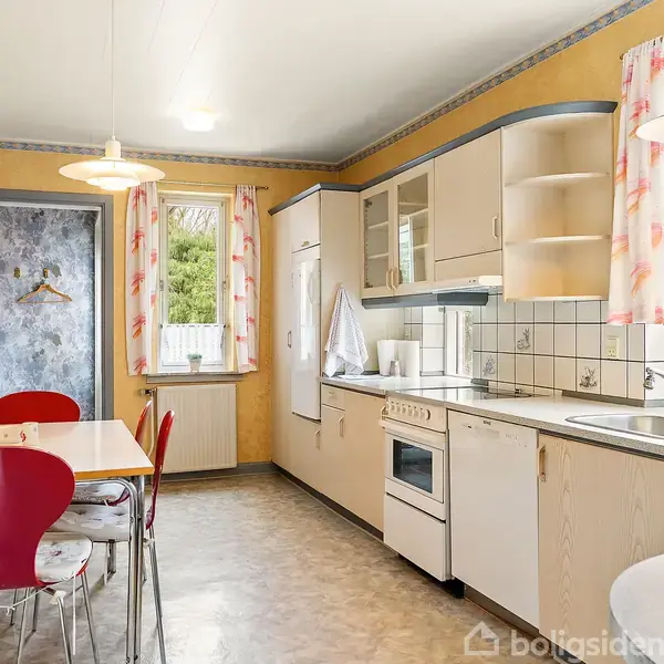 Et køkken med beige skabe og en hvid ovn står i en rummelig, lysegulmalet indretning. Et lille spisebord med røde stole er placeret ved vinduet med blomstrede gardiner.