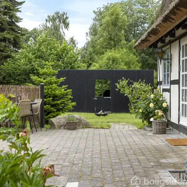 Hus med hvid facade og stråtag står ved en stenbelagt terrasse omkranset af blomster og grønne træer. Et sort træhegn med åbning ses i baggrunden.