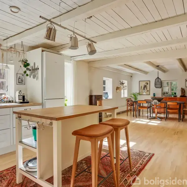 Køkken med en træø og to barstole, omgivet af hvide skabslåger. Lys trægulv og loftsbjælker. Spisebord i baggrunden under en stor lampe, med vinduer giver naturligt lys.