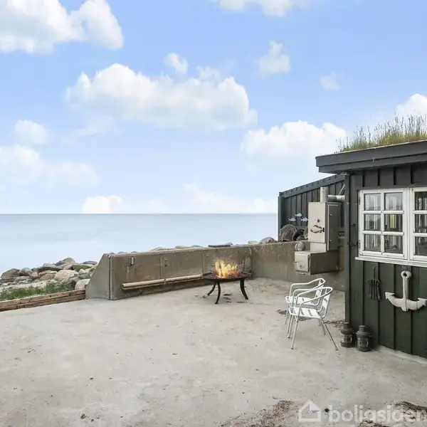 En lille terrasse med en sortmalet hytte og ildsted. Hytten har græs på taget. Baggrunden viser havudsigt og let skyet himmel. Skyllemiddelkanne hænger ved vinduet.