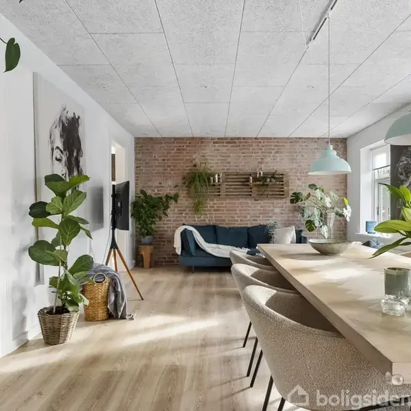 En moderne dagligstue med bord og stole til venstre, planter og kunst på væggen. Baggrunden viser en sofa og dekorative elementer foran en rå murstensvæg.