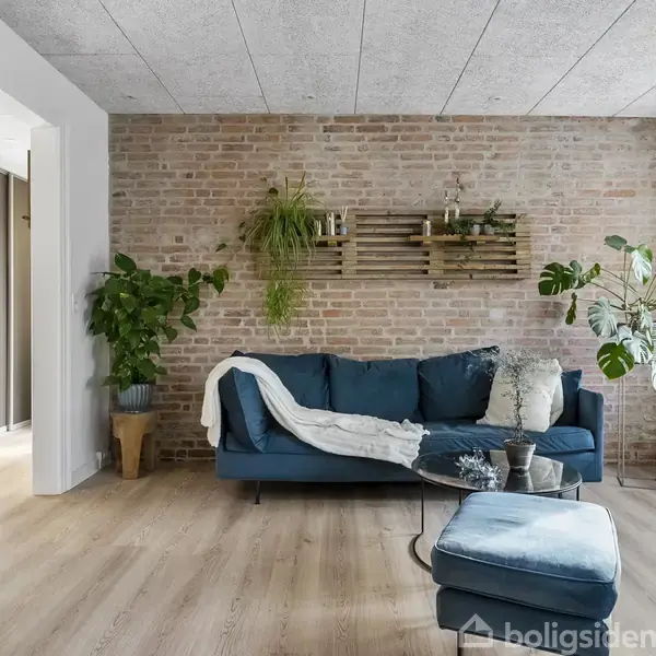Blå sofa står mod en murstensvæg med hængende planter; et blødt tæppe ligger over sofaens armlæn. Et moderne fjernsyn på stativ står til venstre på et lyst trægulv.
