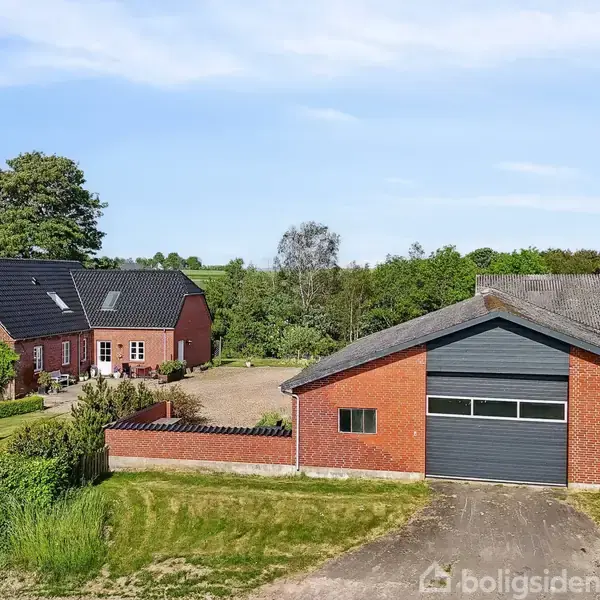 To bygninger i røde mursten står stille i en landlig have, omgivet af græsplæner og træer. En større bygning har en garageport, mens den anden ligner et hus med sorte tage.