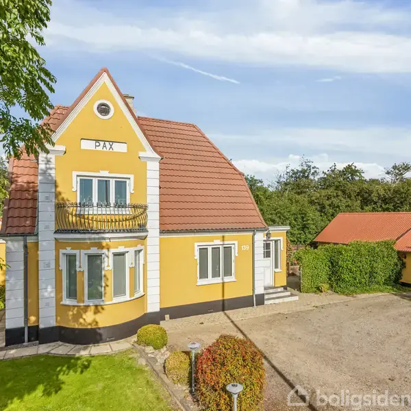 Gul to-etagers villa med teksten "PAX" øverst, byder på buet balkon og rødt tegltag. Omringet af en frodig have og grusbelagt gårdsplads, med rødt tag i baggrunden.
