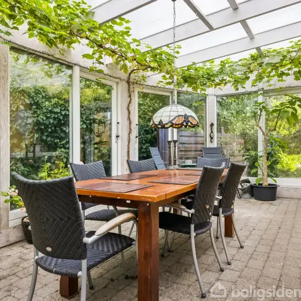 Lang spisebord med flettede stole står i en glaskonservatorie med grønne planter og vinranker, der hænger fra loftet. Udsigt til frodig have gennem store vinduespartier.