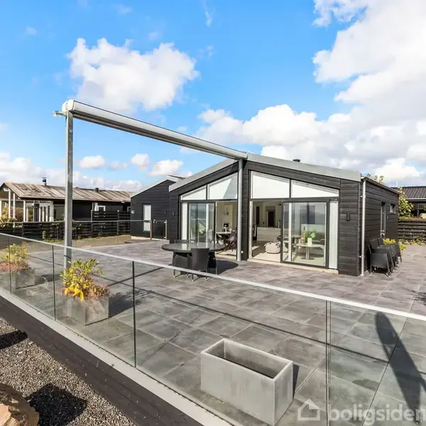 Sort, moderne hus med stor terrasse dekoreret med plantekasser og sten. Terrassen har glasrækværk, og huset har store vinduer. Omgivet af andre huse under en blå himmel med skyer.