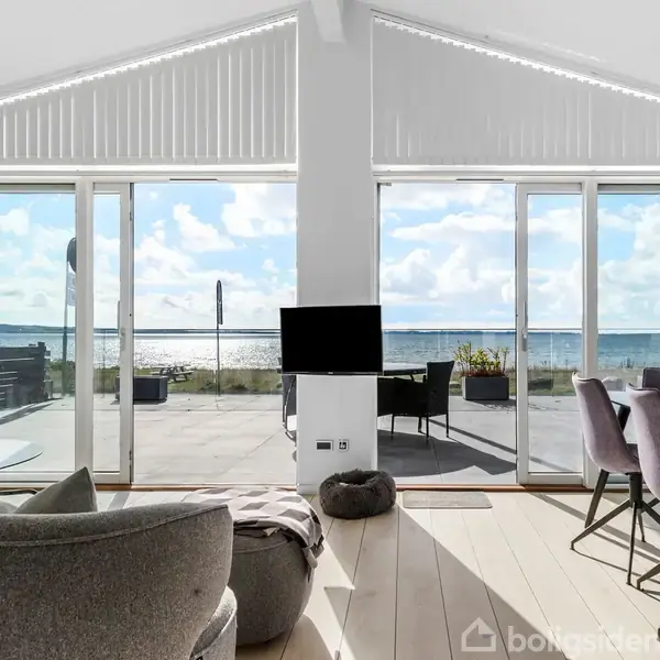 Stuen har en moderne indretning med en grå sofa, spisebord og stole. Store vinduer giver udsigt til en solrig terrasse med havudsigt. En lille sort tv-skærm er midtfor.