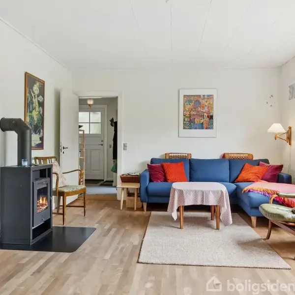 En stue med en blå sofa og farverige puder står centralt. En brændeovn ved siden af er tændt. Rummet har trægulv, lyse vægge, et gulvtæppe og billeder på væggene.