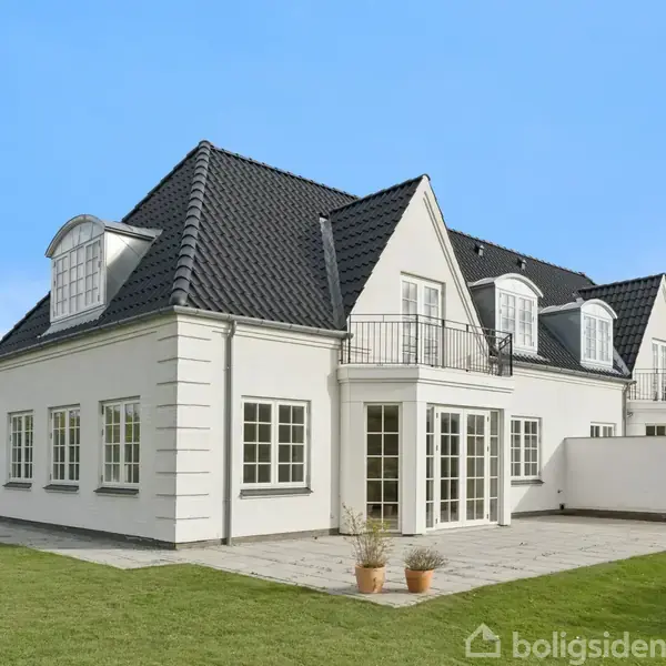 Et stort, hvidt hus med sort tag står i sollys. Det har flere store vinduer og en balkon. Huset ligger i en have med en græsplæne og en flisebelagt terrasse.