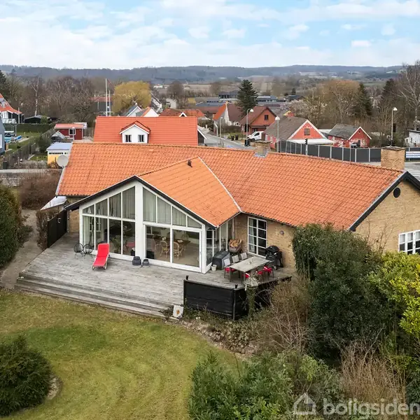 Et hus med rød tegltag og store vinduespartier står stille på en græsplæne med haver. Omgivet af flere andre huse i en rolig nabolagssammenhæng.