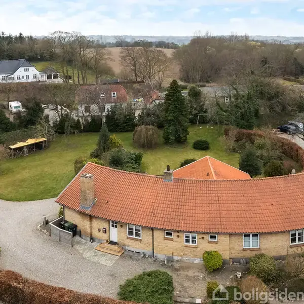 Et rundt, gult murstenshus med rødt tegltag står på en grusvej i et grønt haveområde, omgivet af træer og buske, med andre huse og åbne marker i baggrunden.