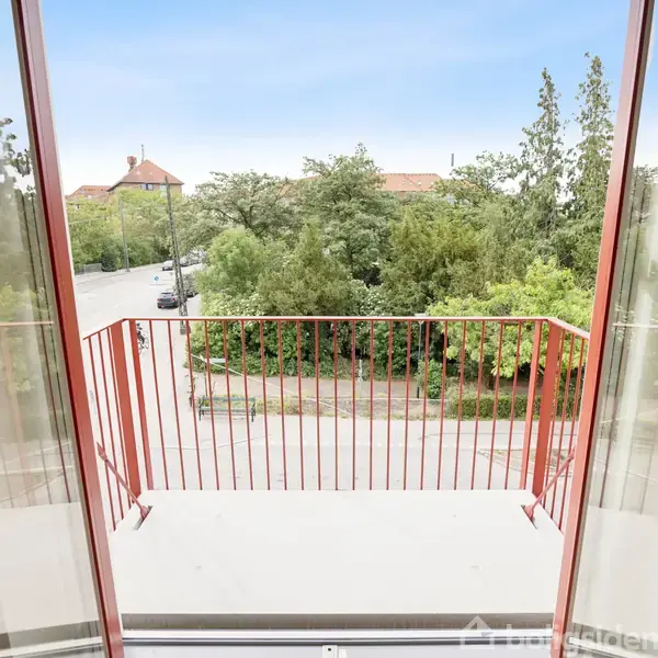 Åben altandør med rødt rækværk, vender ud mod en grøn bypark med træer og en delvist synlig vej i forgrunden.