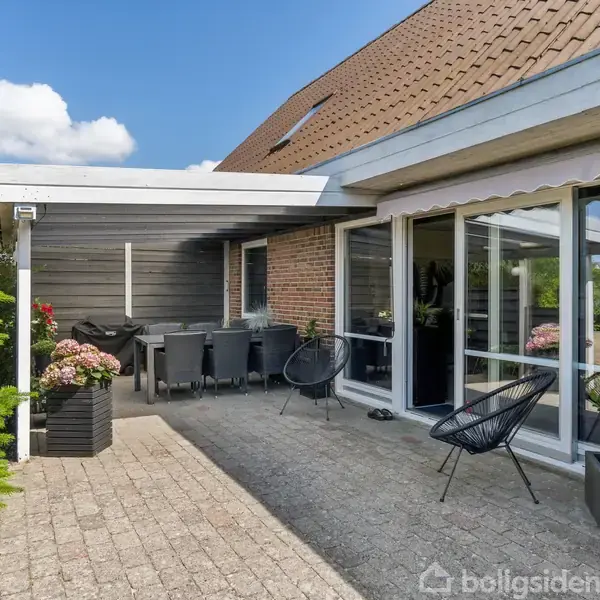 En møbleret terrasse med spisebord og stole under halvtag. Omgivet af murstensvægge og store glasvinduer. Planter i potter placeres langs kanterne.