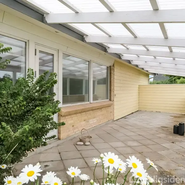 Terrasse med flisebelagt gulv under et hvidt halvtag. Til venstre er der buske og vinduer ind til et hus. I forgrunden blomstrer hvid marguerit i haven.