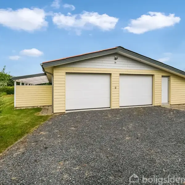 Gul garagebygning med to lukkede garageporte og en dør, placeret på en grusbelagt indkørsel omgivet af græs og buske under en blå himmel med skyer.