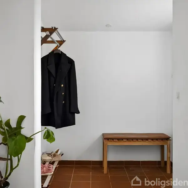 En frakke hænger på en knagerække i en entré med en hvid væg. En bænk står ved siden af, og en plante står ved en væg. Terrazzofliser på gulvet.