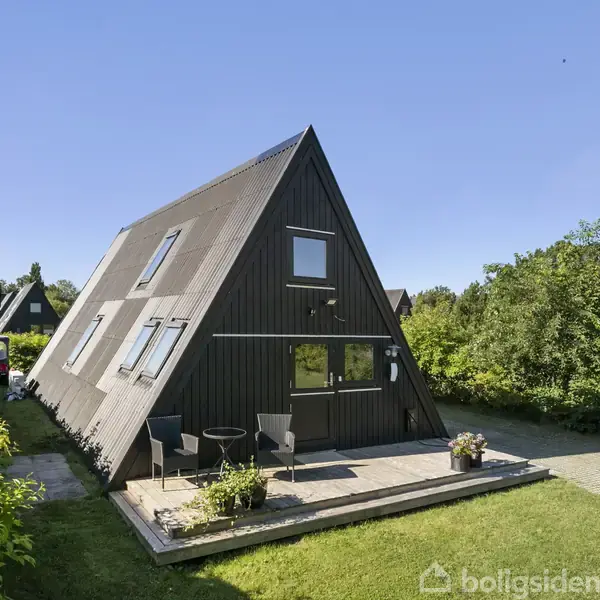 Et A-formet hus står i et grønt, solrigt område. Huset har store vinduer og en terrasse med stole. Naboer kan ses omkranset af frodig vegetation.