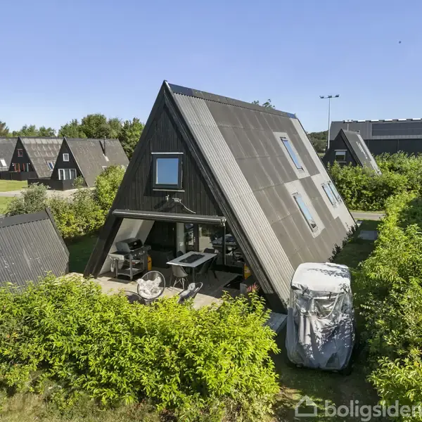 A-formet sommerhus står i en frodig have omgivet af grønne buske. Huset er sort med store vinduer, og der er en terrasse med havemøbler udenfor. Flere lignende huse ses i baggrunden.