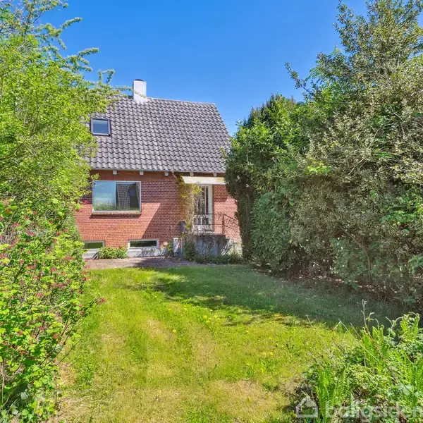 Et murstenshus med skråt tag står omgivet af frodige grønne buske og træer. Foran huset er en velplejet græsplæne under en klar blå himmel.