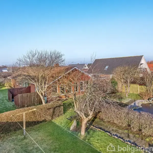 Rødt hus omgivet af en grøn have med træer og hække. En dam med sten ligger til højre. Naboens tag ses i baggrunden under en klar blå himmel.