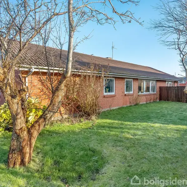 En rød murstensbygning med skråt tag står stille i en have med grøn græsplæne og nøgne træer. En parkeret bil skimtes i baggrunden.