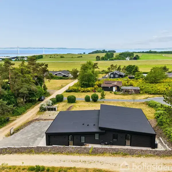 Sort hus med fladt tag står stille omgivet af grønne marker og træer, med udsigt til en kystlinje og vindmøller i baggrunden.