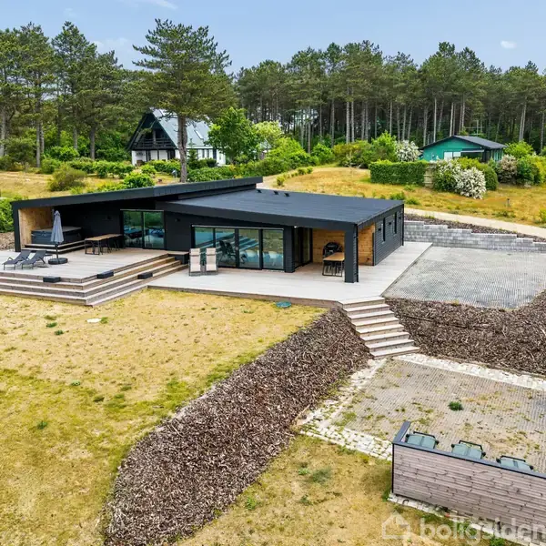 Et moderne, en-etagers hus med store vinduer ligger i en skovklædt område. Omkring huset er der en fri terrasse med en parasol og en græsbelagt gård.