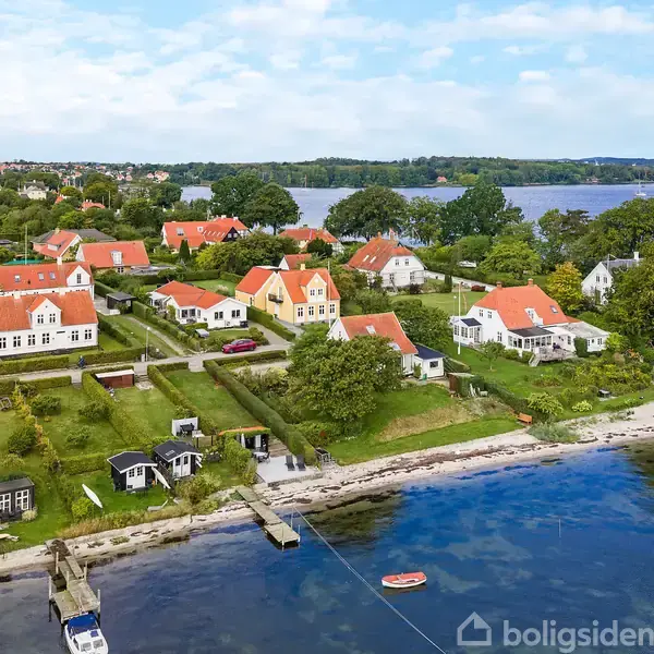 Række af kysthuse med røde tage står stille langs en grøn have ved vandet. Der er både og en lille bro i det klare vand.