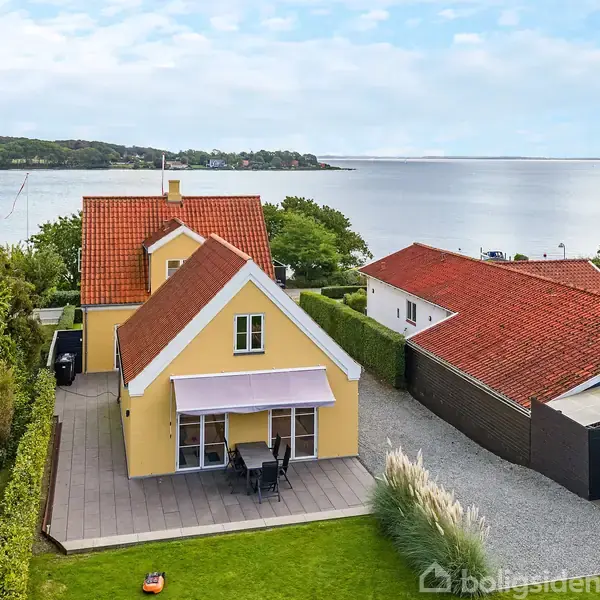 Gul villa med rødt tegltag; har terrasse og havemøbler foran. Havudsigt med kystlinje og skov i baggrunden. Nabo bygninger flankerer huset, omgivet af græsplæne og buske.