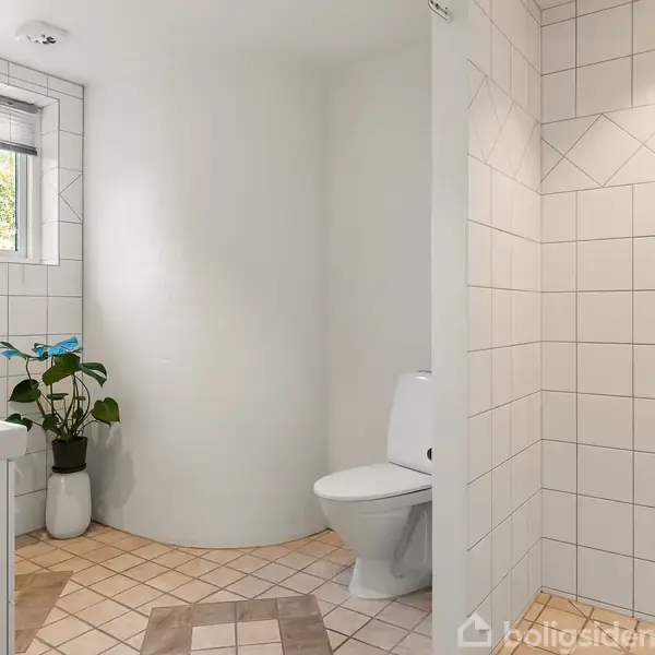 Badeværelse med hvide fliser. Toilet og håndvask er tilstede. En plante står i hjørnet, og et vindue med persienne giver lysindfald. Bruseområdet er afgrænset til højre.