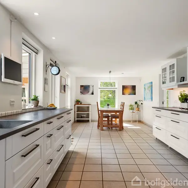 Et moderne køkken har hvidt inventar og sorte bordplader. Et spisebord med stole står i baggrunden. Stort vindue og naturligt lys fylder rummet, dekoreret med planter og kunstværker.