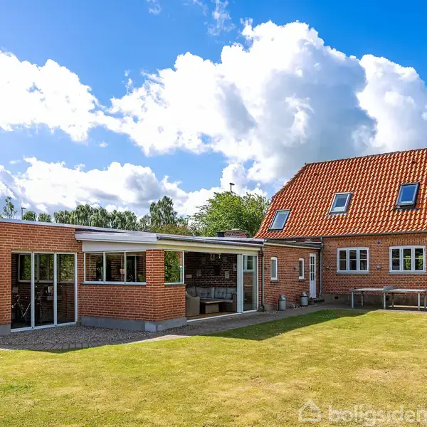 Rødstenshus med rødt tegltag i solrigt vejr, omgivet af en velplejet græsplæne og blå himmel med skyer. Der ses store vinduespartier og en terrasse til højre.