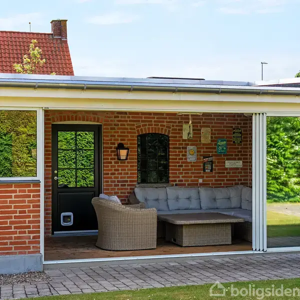 En indglasset veranda med en flettestol og sofa. Verandaen er tilknyttet et hus med røde mursten, omkranset af en grøn have og hække.