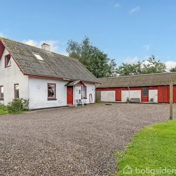 Et hvidt hus med rødt tag og kvist ligger på en grusbelagt gårdsplads, omgivet af grønne træer og en græsplæne. 휴먼 sidder ved indgangen.