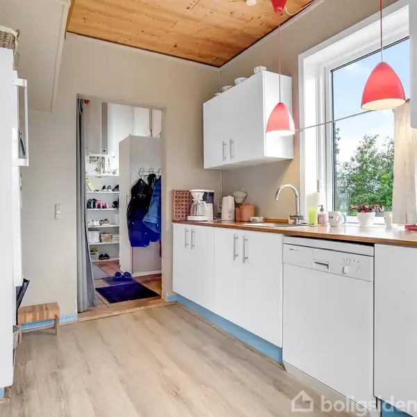 Køkken med hvide skabe og træbordplade under røde hængelamper. Vindue giver udsigt til træer. Til højre ses en ovn, til venstre et åbent dørparti mod en gang.