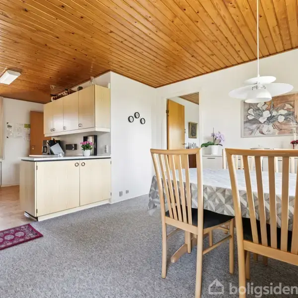Et køkken-alrum med spisebord dækket af dug og træstole. Rummet har trægulv i køkkenområdet, gråt tæppe ved spisepladsen og træloft. En hvid loftlampe hænger over bordet.