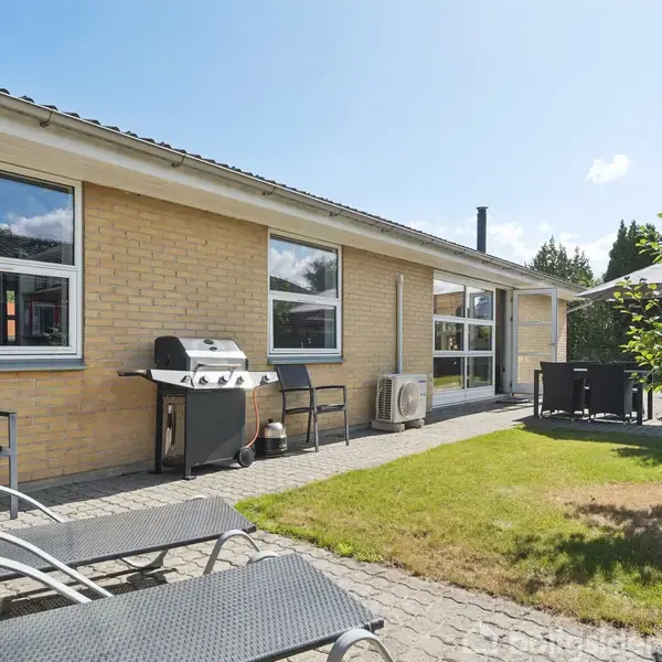 En gul murstensbygning med store vinduer; foran står to liggestole og en grill på en flisebelagt terrasse. I baggrunden ses græsplæne, et lille træ og nogle havemøbler.