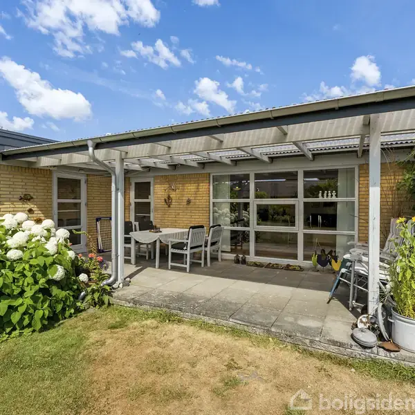 Husets terrasse er dækket af et halvtag med havemøbler, omgivet af frodige planter, herunder hortensiaer. Konteksten er en velplejet have under en klar, blå himmel.