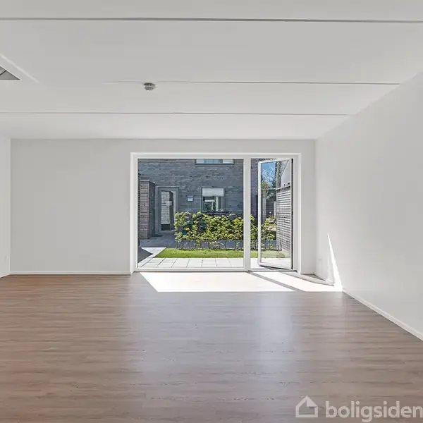 Stue med trægulv og åbent område fører ud til en terrasse med græsplæne gennem store skydedøre. Hvide vægge omkranser rummet, og trapper er synlige til venstre.