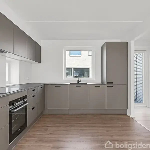 Køkkenet har grå skabe og skuffer, en indbygget ovn og kogeplade, alt på et lyst trægulv. Et vindue tilføjer lys, ved siden af indgangen til en anden del af huset.