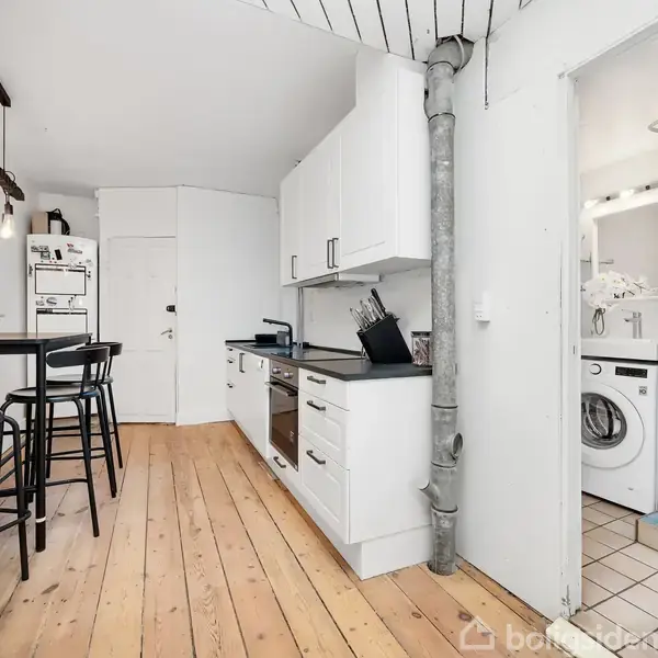 Køkken med hvidt skab og ovn langs væggen, trægulv og spisebord med høje stole. Til højre ses et tilstødende rum med vaskemaskine og spejl.