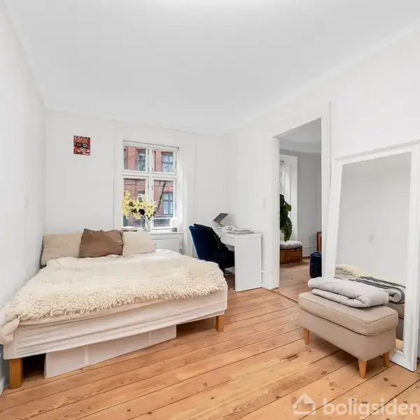 En seng med hvide sengetæpper står i et lyst rum med trægulv. En stor spejl hviler mod væggen ved siden af en puf. Et vindue bagved viser en murstensbygning.