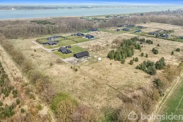 Grå bygning står stille i landsbyområde omgivet af åbne marker og spredt bevoksning, med kystlinje og skov i baggrunden. Boligområde med flere huse i nærheden.