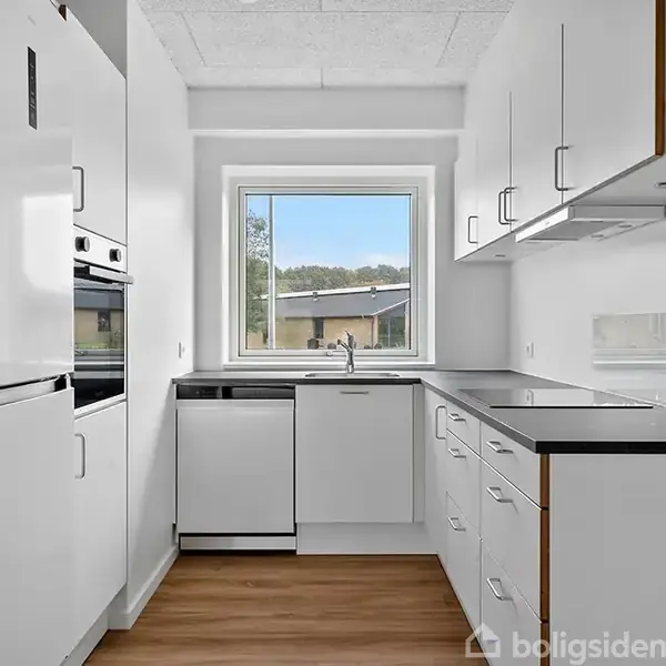 Et hvidt, moderne køkken med integrerede apparater og sorte bordplader. Køkkenet har et vindue, der giver udsigt til et udendørs område, omgivet af en lys trælaminatgulv.