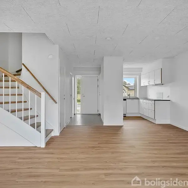 Trappe med trægelænder fører opad; omgivet af et rummeligt, lyst rum med lys trælaminatgulv. Køkken i baggrunden har hvide skabe og en stor vindue; minimal, moderne indretning.